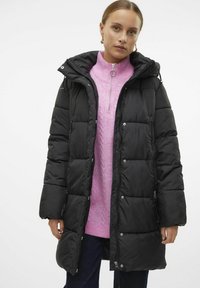 Svart puffjacka med en quiltad design, dragkedjefickor och huva, bärs över en ljusrosa stickad tröja.