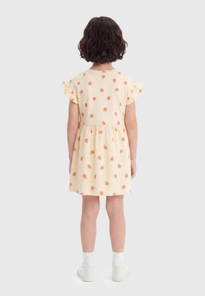 Kind met kort krullend haar, een gele jurk met oranje fruitprint, witte sokken en witte sneakers, staand met de rug naar de kijker.