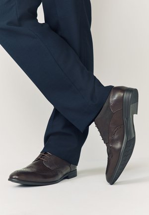 Homme portant des chaussures habillées en cuir marron foncé et un pantalon formel bleu marine, debout avec un pied croisé sur l'autre.
