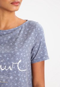 Camiseta gris de manga corta con un patrón floral en azul claro. Presenta texto en script blanco y un escote relajado. Tejido suave y casual.