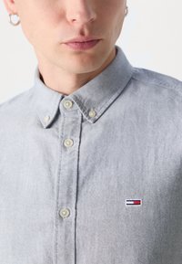 Ανοιχτό γκρι πουκάμισο με κουμπιά, φτιαγμένο από βαμβακερό μείγμα υφάσματος; διαθέτει ένα μικρό κεντημένο λογότυπο στην αριστερή πλευρά του στήθους και κουμπιά στην πλάκα.