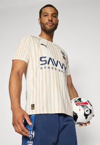 Beidzas īsu piedurkņu futbols krekliņš ar vertikālām svītrām, melns Puma logo un teksts "SAVVY GAMES GROUP". Spēlētājs tur melnbaltu futbolu.