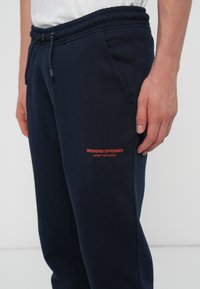 Marineblå joggingbukser med snoretræk i taljen, sidelommer og et orange trykt logo, der står "WEEKEND OFFENDER" og "ADMIT NOTHING."