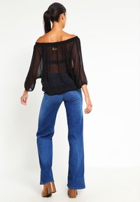 Kobieta z ciemnym kucykiem, ubrana w czarną przezroczystą bluzkę z odsłoniętymi ramionami, niebieskie dżinsy i brązowe buty na obcasie, stojąca tyłem na białym tle.