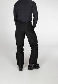Personne portant un pantalon de ski noir et des chaussures de ski noires, debout de dos face au spectateur sur un fond uni.