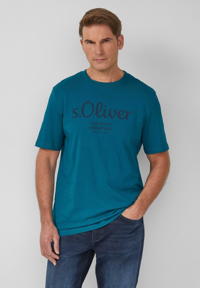 Türauschnitt T-Shirt in Türkis mit kurzen Ärmeln und Rundhalsausschnitt, versehen mit dunkelblauem Text "s.Oliver Elevated Essentials Since 1969", kombiniert mit dunkelblauen Jeans.