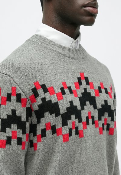 Άνδρας που φοράει ένα γκρι πλεκτό πουλόβερ με κόκκινες και μαύρες γεωμετρικές σχέσεις, πάνω από ένα λευκό πουκάμισο με γιακά.