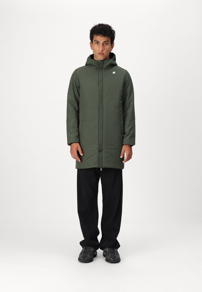 Veste à capuche verte avec fermeture éclair à l'avant, présentant une texture lisse et un logo sur la poitrine. Portée sur un pantalon noir et des chaussures de sport.
