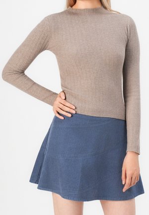 Vrouw draagt een taupe geribd longsleeve top en een blauwe uitlopende corduroy minirok, hand rustend op de heup.