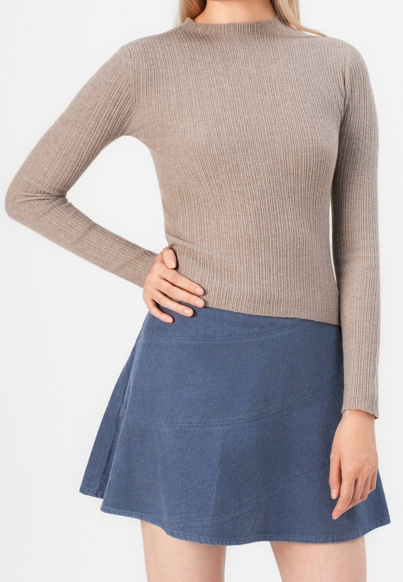 Femme portant un haut à manches longues côtelé couleur taupe et une mini-jupe évasée en velours côtelé bleu, main reposant sur la hanche.