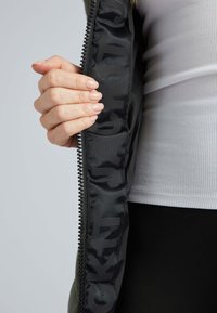 Veste noire avec une fermeture éclair sur le devant, présentant un matériau extérieur lisse et des lettres audacieuses à l'intérieur. Une main saisit la fermeture sur la couture.