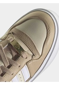 Beige lädersneakers med texturerade paneler, en slät gräddfärgad tåkappa, guldaccenter och vitt randigt detaljer.