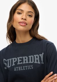 T-shirt à manches longues bleu marine avec le texte SUPERDRY ATHLETICS en gris. Tissu en coton avec un col ras du cou et une coupe décontractée.