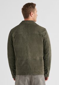 Grüne Wildlederjacke mit einem Kragen, rückseitigen Nahtdetails und einem knöpfbaren Ärmel. Der Stoff hat ein weiches, strukturiertes Finish.