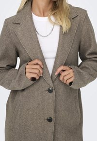 Manteau marron en mélange de laine avec un col à revers, des boutons noirs et des poches latérales. Texture lisse, design mi-long, porté par-dessus un haut blanc.
