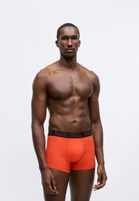 Red cotton boxer briefs with a black logo waistband, fitted design, and smooth texture, displayed on a model against a plain background.