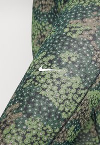 Primo piano di leggings Nike a motivo floreale verde con il logo Nike swoosh bianco sulla coscia e cuciture nere visibili.