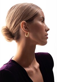 Złota kolczyk w kształcie obręczy o skręconym wzorze, prezentująca lśniącą powierzchnię. Modelka ma elegancki kok i nosi głęboko fioletowy strój.