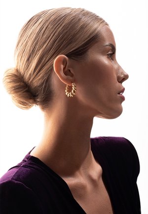 Złota kolczyk w kształcie obręczy o skręconym wzorze, prezentująca lśniącą powierzchnię. Modelka ma elegancki kok i nosi głęboko fioletowy strój.