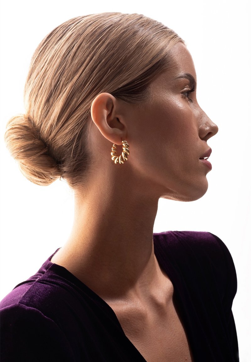 Złota kolczyk w kształcie obręczy o skręconym wzorze, prezentująca lśniącą powierzchnię. Modelka ma elegancki kok i nosi głęboko fioletowy strój.