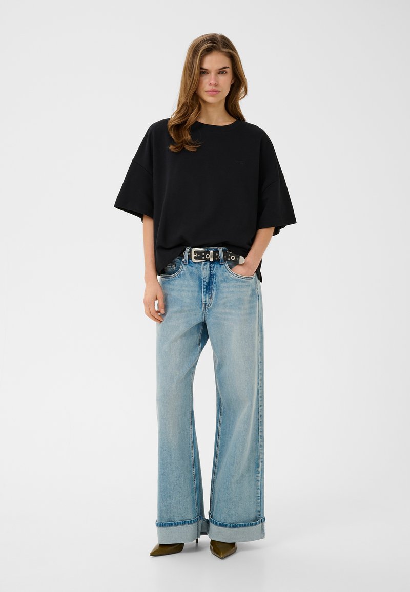 Jeune femme portant un t-shirt noir ample, un jean bleu clair à jambes larges avec revers, une ceinture noire et des talons bruns pointus, debout sur fond blanc.
