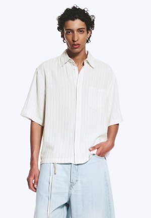 Jeune homme aux cheveux bouclés portant une chemise blanche ample à manches courtes boutonnée et un jean bleu clair à jambes larges, une main dans la poche.