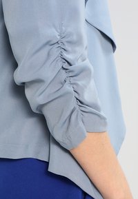 Ljusblå blus med samlad detalj vid ärmslutet, i ett mjukt och slätt tyg och med en avslappnad passform.
