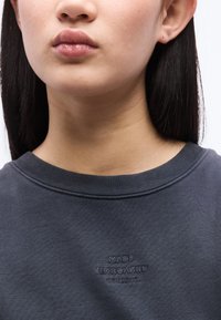 Femme aux cheveux raides et foncés portant un sweat-shirt gris foncé à col rond avec "Mads Nørgaard Copenhagen" brodé sur la poitrine.