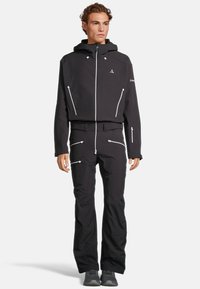 Set di giacca e pantaloni neri per esterni realizzato in materiale softshell. Presenta cerniere, cappuccio regolabile e numerose tasche con cerniera.