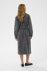 Grå denimklänning med bälte, långa ärmar, midikalvlängd, synliga sömmar och en fast, texturerad yta. Bärs med svarta ballerinaskor.