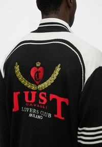 Čierna pletená bunda so základom vyšívanej značky "JUST CAVALLI" v červenej a zlatej farbe, srdcom, vavrínovými listami a bielymi pruhmi na rukávoch.