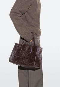 Persona con maglione marrone e pantaloni grigi che tiene una borsa in pelle marrone scuro con texture, afferrandola per i manici lungo il fianco.