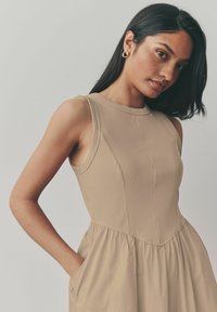Sømløs beige kjole med ribbet overdel og samlede skørt, med sidelommer og høj halsudskæring. Glat tekstur, minimalt design.