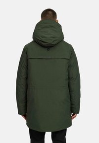 Man in een donkergroen gewatteerd winterparka met capuchon, gezien vanuit de achterkant tegen een effen lichte achtergrond.