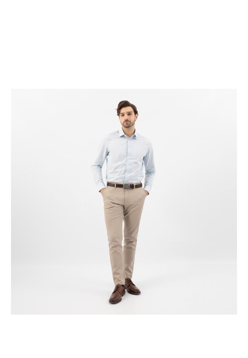 Mężczyzna ubrany w jasnoniebieską koszulę na guziki, beżowe spodnie, brązowy pasek i brązowe buty wizytowe stoi z rękami w kieszeniach na białym tle.
