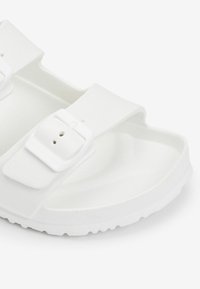 Sandale blanche dotée de deux sangles réglables avec boucles, fabriquée à partir d'un matériau texturé, avec un fond de chaussure contouré et une semelle extérieure flexible.