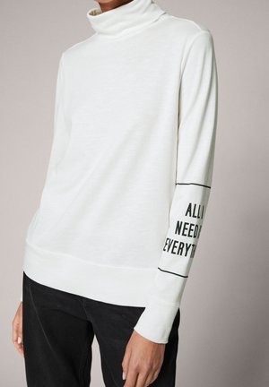 Biała bluzka z długim rękawem i golfem z czarnym napisem „ALL I NEED IS EVERYTHING” na prawej przedramieniu, noszona z czarnymi spodniami.