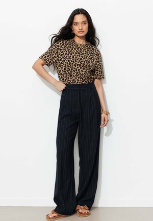 Femme aux longs cheveux noirs portant un t-shirt imprimé léopard, un pantalon large noir à rayures taille haute et des sandales couleur beige, debout contre un mur blanc.