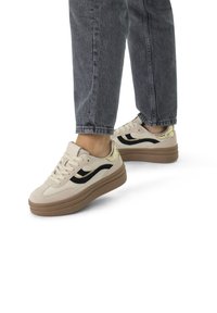 Beige suede sneakers with a black wave design and gold accents, featuring a chunky brown sole and traditional lacing system.