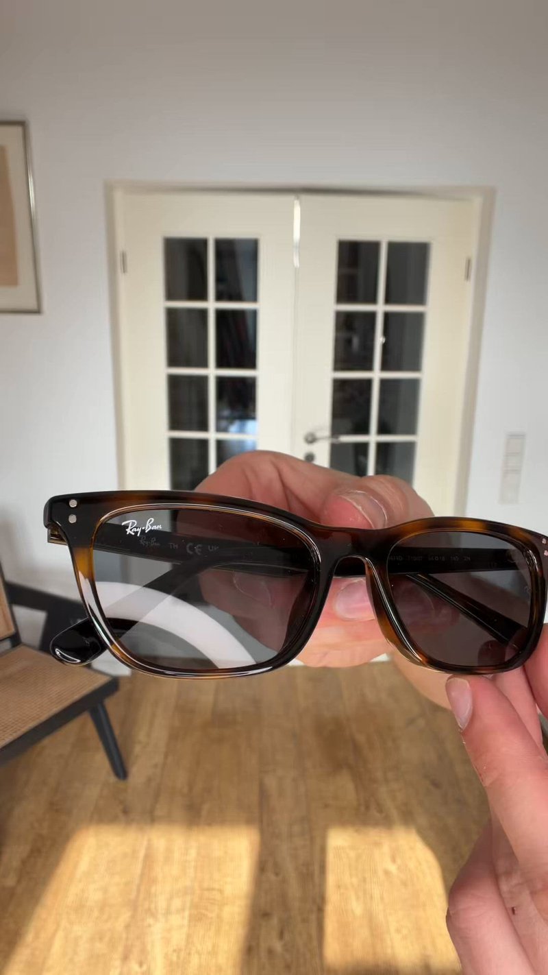 Ръка държи правоъгълни очила Ray-Ban с костенурков десен, с прозрачни и тонирани лещи, на закрито до бели двукрили врати.