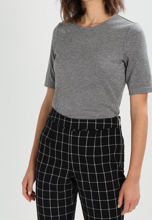 Femme portant un haut gris moulant à manches courtes avec un motif à petits carreaux fins et un pantalon noir avec motif à grandes fenêtres blanches, debout devant un fond uni.