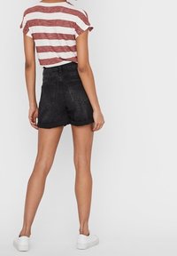 Person klädd i en röd-och-vit randig t-shirt, svarta denimshorts med uppvikt fåll, och vita sneakers, står med ryggen vänd mot kameran.