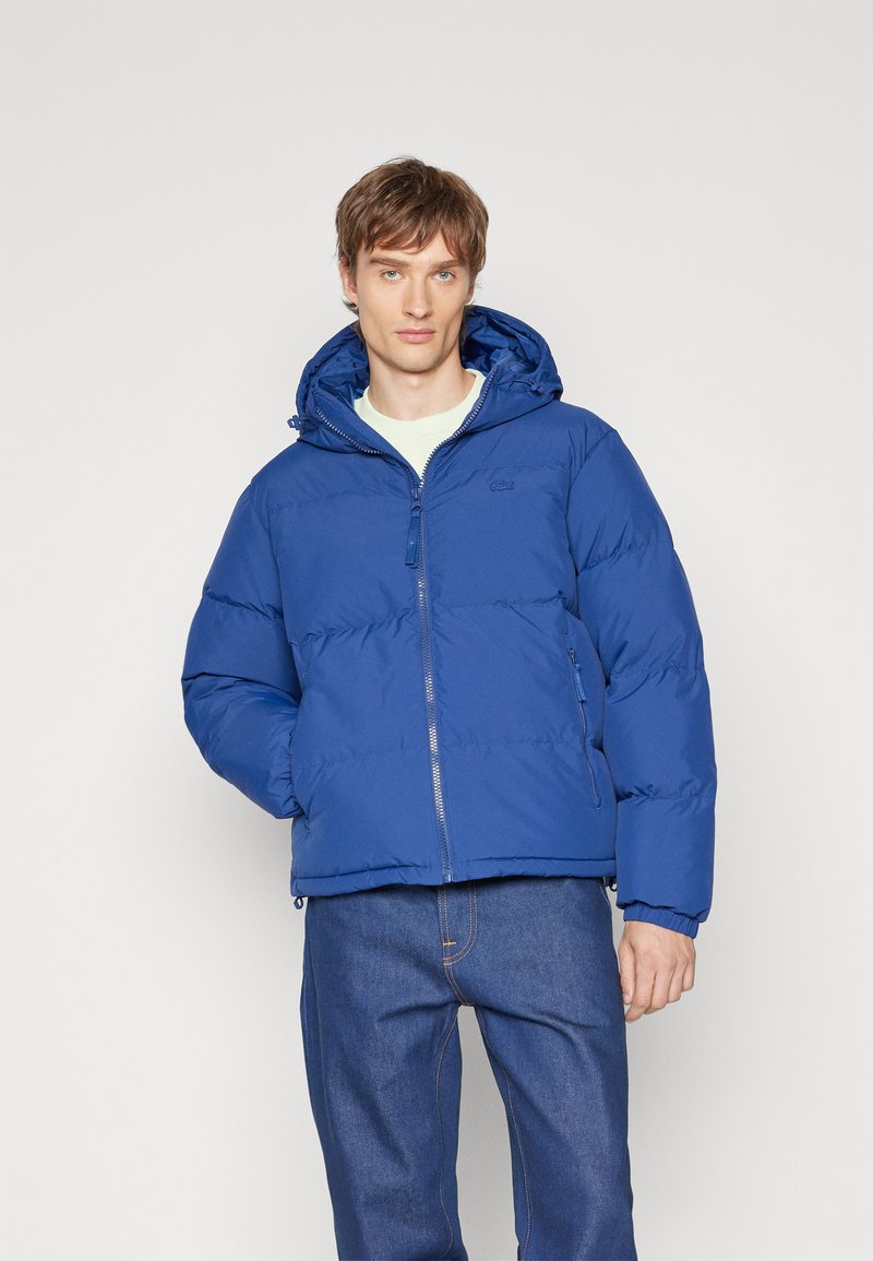 Chaqueta acolchada azul con capucha y diseño de acolchado, que cuenta con una cremallera en la parte delantera y bolsillos laterales, combinada con pantalones de mezclilla azules.