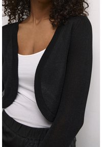 Schwarze, kurze Strickjacke mit strukturiertem Strick und offenem Design, getragen über einem weißen Oberteil; verfügt über lange Ärmel und schimmerndes Material.
