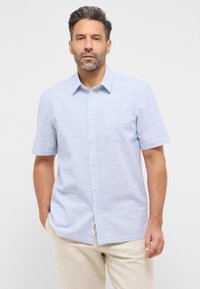 Jasnoniebieska koszula z krótkim rękawem zapinana na guziki, z delikatną teksturą, kieszonką na piersi i beżowymi guzikami. Noszona z jasnymi spodniami.