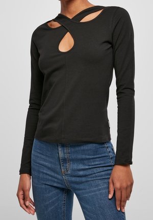 Femme portant un haut noir à manches longues avec deux découpes près de la clavicule, associé à un jean bleu sur un fond uni.