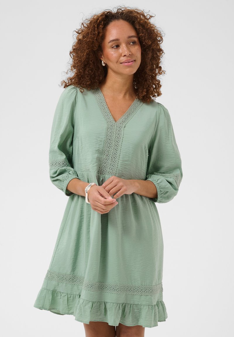 Femme aux cheveux bouclés portant une robe vert clair à col en V avec des détails en dentelle, debout, les mains doucement jointes.