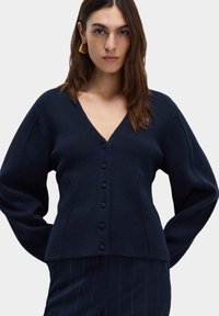 Granatowy sweter robiony na drutach z dekoltem w serek, z zapięciem na guziki i długimi rękawami, charakteryzujący się miękką fakturą i dopasowanym krojem.