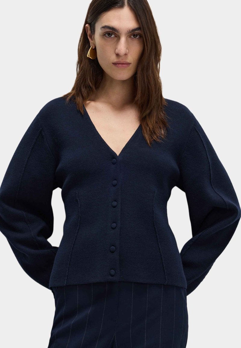 Granatowy sweter robiony na drutach z dekoltem w serek, z zapięciem na guziki i długimi rękawami, charakteryzujący się miękką fakturą i dopasowanym krojem.