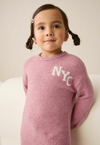 Junge mit geflochtenen Zöpfen und Haarclip, der einen pinken Pullover mit dem Schriftzug "NYC" trägt und vor einem hellen Hintergrund steht.